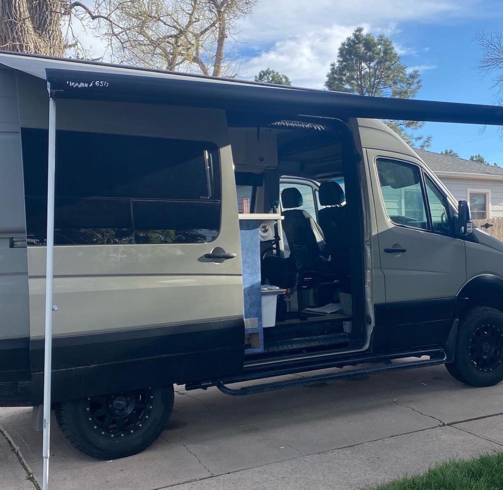 A 4x4 Mercedes-Benz Sprinter camper van is parked with the slider door open and the awning deployed.