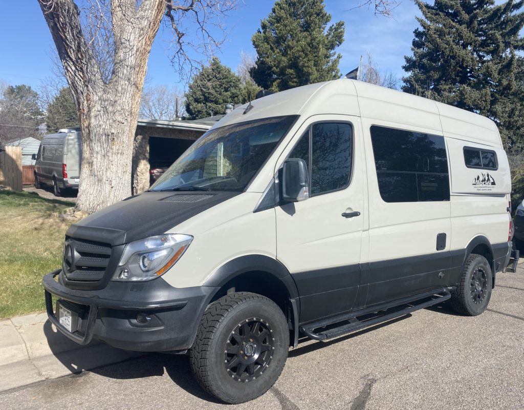 A 4x4 Mercedes-Benz Sprinter camper van is parked next to a tree.